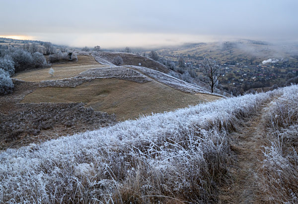 Winter coming. Picturesque pre sunrise scene above late autumn mountain countryside with hoarfrost on grasses, trees, slopes. Peaceful sunlight rays from cloudy sky. Ukraine, Carpathian Mountains.