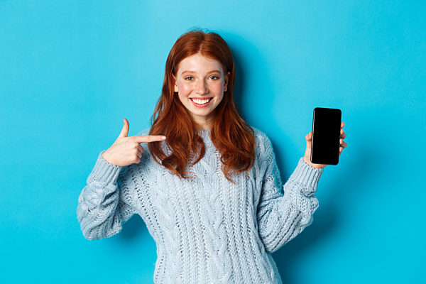 Satisfied redhead girl pointing at phone screen, showing smartphone app or online promo and smiling, standing in sweater against blue background