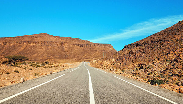 Straight asphalt road , small Atlas mountains and low bushes both sides, clear sky above - typical landscape in Southern Morocco