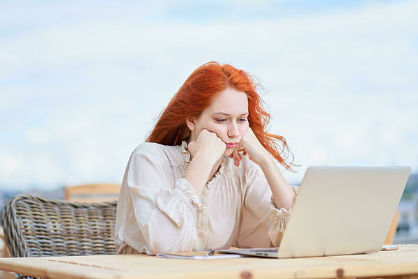 Bored woman sitting in cafe on street and remotely working on laptop. Redhaired female