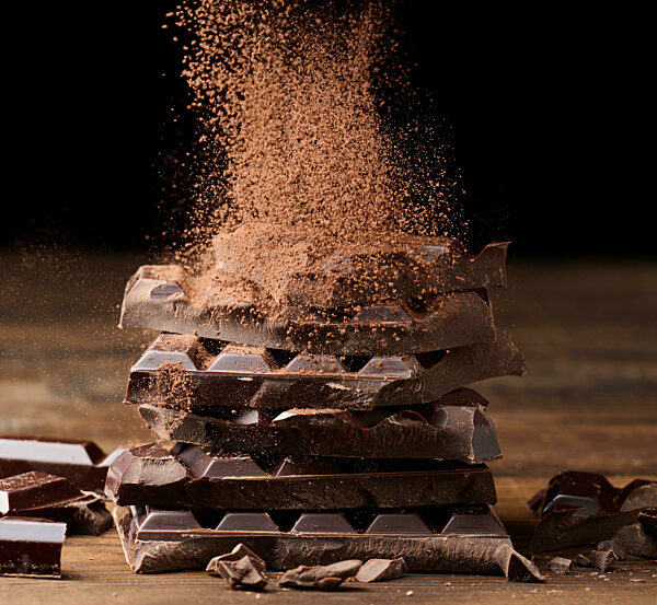 cocoa powder sprinkles on a stack of broken dark chocolate on a wooden table, black background