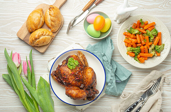 Easter food on white rustic table: colored eggs, roasted chicken, vegetables, buns