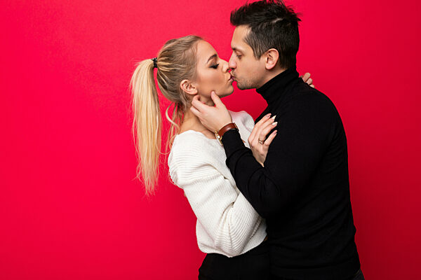 Photo of a beautiful young blonde woman white knitted sweater and an adult brunet man with stubble in a black sweater who are kissing isolated on a pink background with free space for text. Love concept