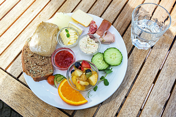 Breakfast or lunch plate with bread rolls, cheese, sausage and fruit on a wooden outdoor table on a sunny day, selected focus