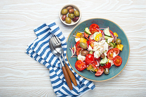 Greek salad with feta cheese, vegetables, olives in blue bowl on white wooden background