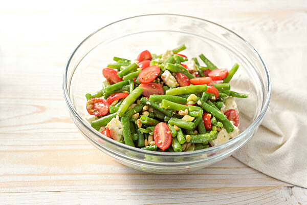 Green bean salad with tomatoes, pine nuts and feta cheese in a glass bowl on a light wooden table, Mediterranean snack or antipasto, copy space, selected focus