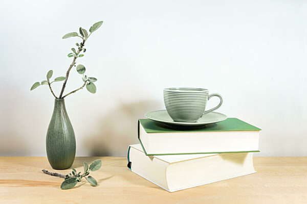 Wooden desk or table with books, a green tea cup and sage twigs in a small ceramic vase against a gray white wall, copy space, selected focus