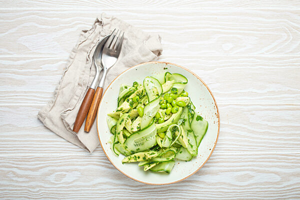 Healthy vegan green avocado salad bowl with sliced cucumbers, edamame beans, olive oil and herbs on ceramic plate top view, white wooden rustic table background
