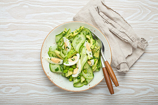 Healthy green avocado salad bowl with boiled eggs, sliced cucumbers, edamame beans, olive oil and herbs on ceramic plate top view, white wooden rustic table background