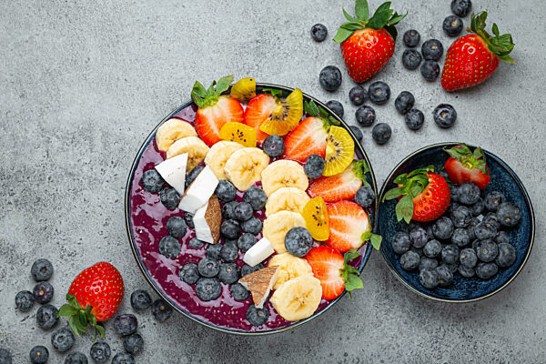 Healthy summer acai smoothie bowl with chia seeds, fresh banana, strawberry, blueberry, cocos, kiwi top view, rustic concrete background with spoon