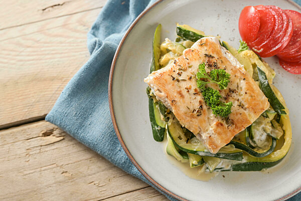 Roasted wild salmon fillet with parsley garnish on creamy zucchini vegetables and fresh tomato on a light plate, healthy meal for a low carb diet, high angle view from above, copy space