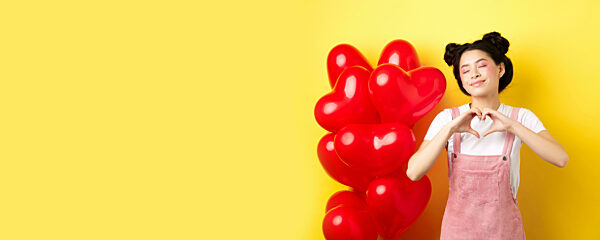 Valentines day concept. Cute asian girl dreaming of romance, close eyes and showing heart gesture, smiling happy, standing near red romantic balloons, yellow background