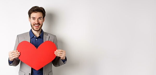 Happy man confessing in love, showing heart cutout and smiling at camera, standing in suit on romantic date with lover, white background