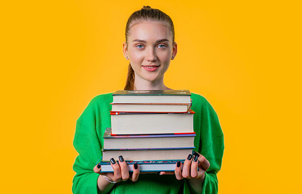 Smiling young student woman with stack of books from library, yellow background