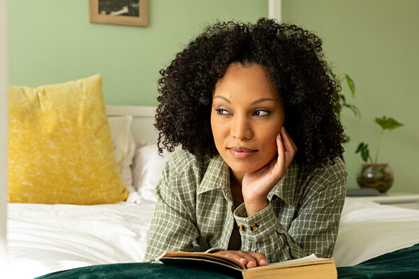 Biracial woman reading book lying on bed in bedroom