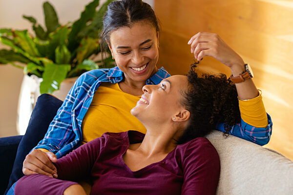 Happy biracial lesbian couple lying on sofa in living room