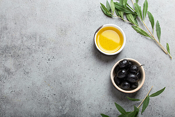 Black olives and extra virgin olive oil in little bowl with olive tree branches top view on grey concrete stone background, space for text