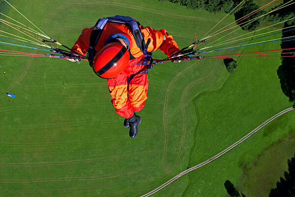 a man in signal red overalls hangs from a paraglider high above the meadows near Lenggries