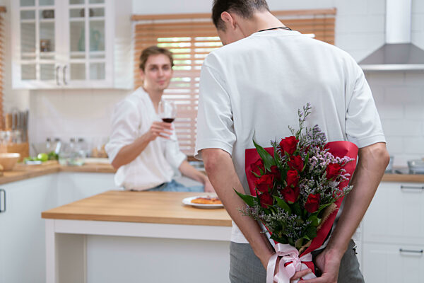 A young LGBT couple celebrates the day of love with a glass of wine in their home kitchen. with a surprise with a rose from a lover
