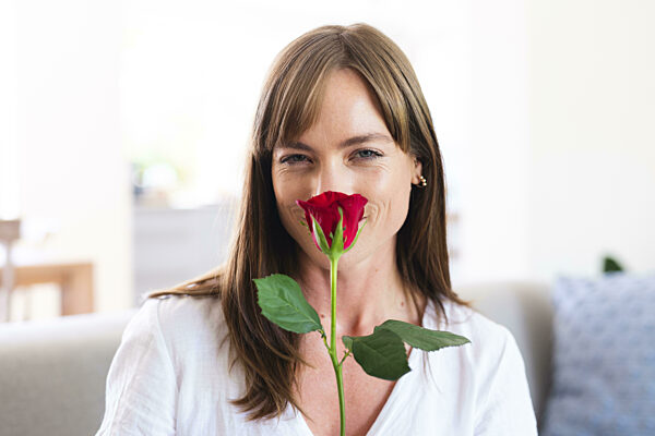 A middle-aged Caucasian woman smiles while holding a red rose close to her face
