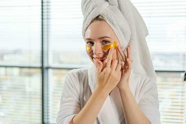 Schönheitsbehandlung mit Augenklappen. Frau mit unter Auge Kollagen Pads. Mädchen mit Patches unter den Augen Handtuch auf dem Kopf bekommt Gesichtsbehandlung. Junge schöne Frau im Bademantel und Handtuch entspannt im Bad zu Hause.