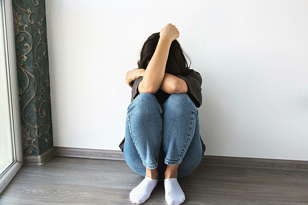 Traurige junge Frau, die auf dem Boden sitzt, die Arme um die Knie geschlungen, ihr Gesicht verbergend, Einsamkeit und Depression ausdrückend