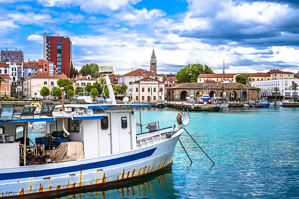 Fischerboot im türkisfarbenen Hafen mit der historischen Stadt Koper und dem wolkenverhangenen Himmel im Hintergrund, Adriaküste von Slowenien