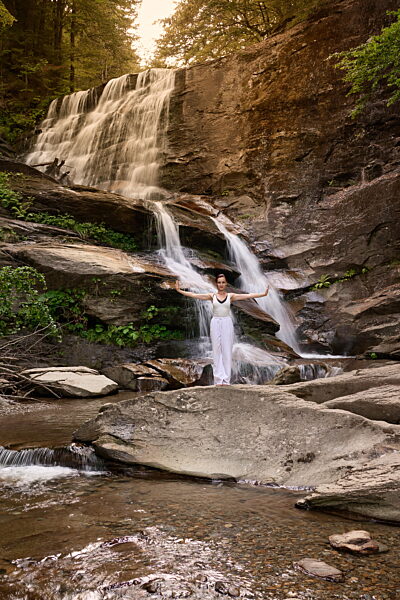 Frau, die in der Nähe eines Wasserfalls und eines Flusses bei Sonnenuntergang langsame Tai-Chi-Bewegungen ausführt und dabei Harmonie, Gleichgewicht und Fitness in der Natur festhält