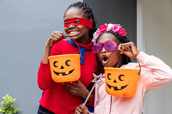 Afroamerikanische Mädchen im Vorschulalter lächeln und jubeln am Hauseingang, während sie Kürbiskörbe in den Händen halten. Jack-O'-Lantern, Kostüme, Duo, Veranda, Halloween, skurril, lebhaft