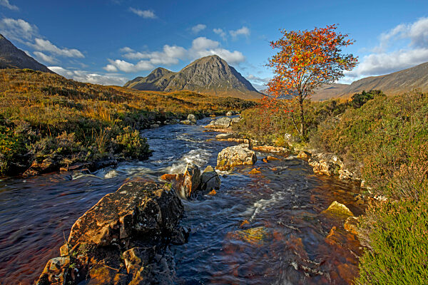 An autumnal view of a stream flowing over moorland towards a mountain.
