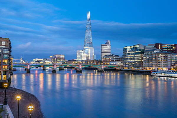 Southwark Bridge and The Shard seen from the riverbank of the River Thames during twilight.