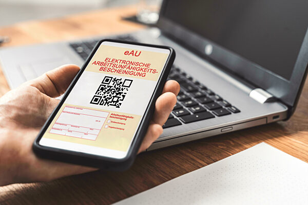 Smartphone mit einer eAU Elektronischen Arbeitsunfähigkeitsbescheinigung in der Hand vor einem Laptop Computer. Elektronische Krankmeldung FOTOMONTAGE