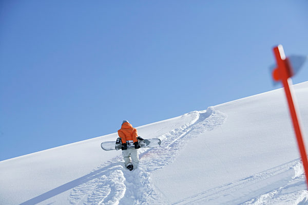Eine Frau laeuft mit einem Snowboard durch Tiefschnee den Berg hoch - Wintersport - Urlaub