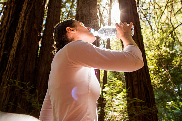 Woman drinking water from bottle in sunny forest, Frau trinkt Wasser aus der Flasche in sonnigem Wald