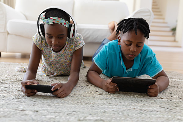 African american brother and sister wearing headphones lying on floor using smartphones