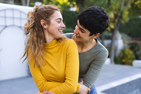 Happy biracial lesbian couple standing on garden terrace embracing and smiling at each other. Love, gay, relationship, togetherness, domestic life and lifestyle, unaltered.