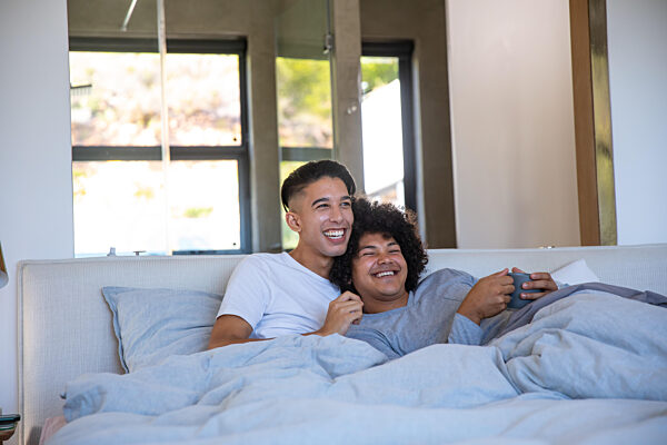 Two men cuddling on bed beneath gray duvet in bright bedroom, holding mug under sunlight glow. Romance, intimacy, comfort, relaxation, modern, cozy, affectionate