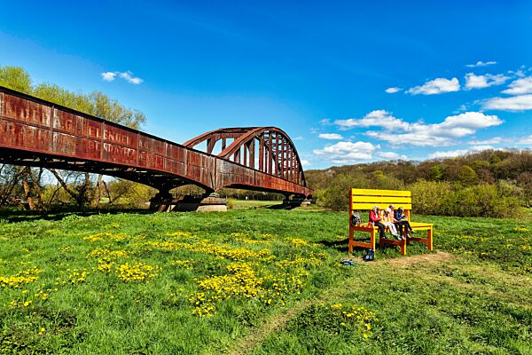 Besucher sitzen auf großer gelber Holzbank vor alter Eisenbahnbrücke, Wiese am Weserufer, sonniges Frühlingswetter, Landesgartenschau 2023, Höxter, Nordrhein-Westfalen, Deutschland, Europa