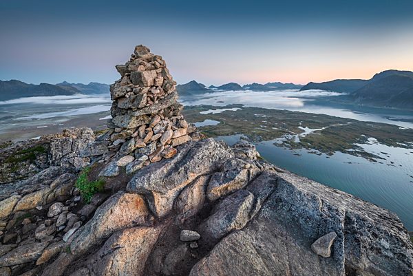 Steinpyramide am Gipfel des Hoven in der Abenddämmerung, hinten Gebirge am Horizont und Fjorde mit Wolkendecke, Gimsøy, Lofoten, Nordland, Norwegen, Europa