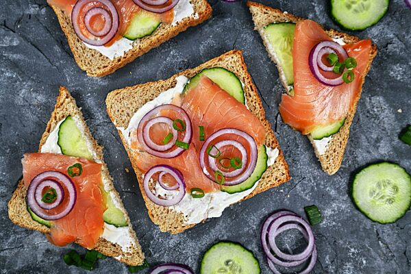 Lachsbrotscheiben aus gesundem Vollkorntoast, belegt mit geräuchertem Lachsfisch, roten Zwiebeln und Frühlingszwiebeln und Gurke auf Meerrettich