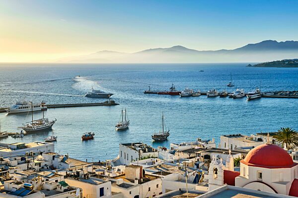 Schöne Aussicht auf Chora, Mykonos, Griechenland bei Sonnenuntergang. Zivilen Hafen im Hafen, Boote, Segelboote, Yachten vertäut durch Steg. Berühmte weiß getünchte Häuser und traditionelle weiße Kirche mit roter Kuppel mit Blick auf den dunstigen H