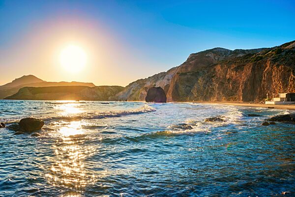 Schöne Landschaft am Strand von Fyriplaka, Milos, Griechenland. Low Sonne über azurblauen Meerwasser, felsigen Ufern, Sandstrand, dunstigen fernen Felsen. Bunte Sonne und Himmel, Sonnenfackeln auf Wellen