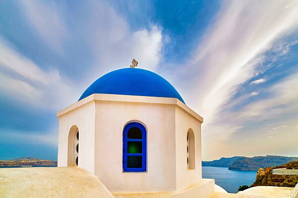 Weiße griechisch orthodoxe Kirche und ihre blaue Kuppel mit Kreuz gegen dramatischen blauen Himmel und Wolken, Santorini Insel, Griechenland. Schöne Landschaft, Meer Gewässer, Inseln, Hafen, Klippen, Reiseziele im Mittelmeer Kreuzfahrten, touristische A
